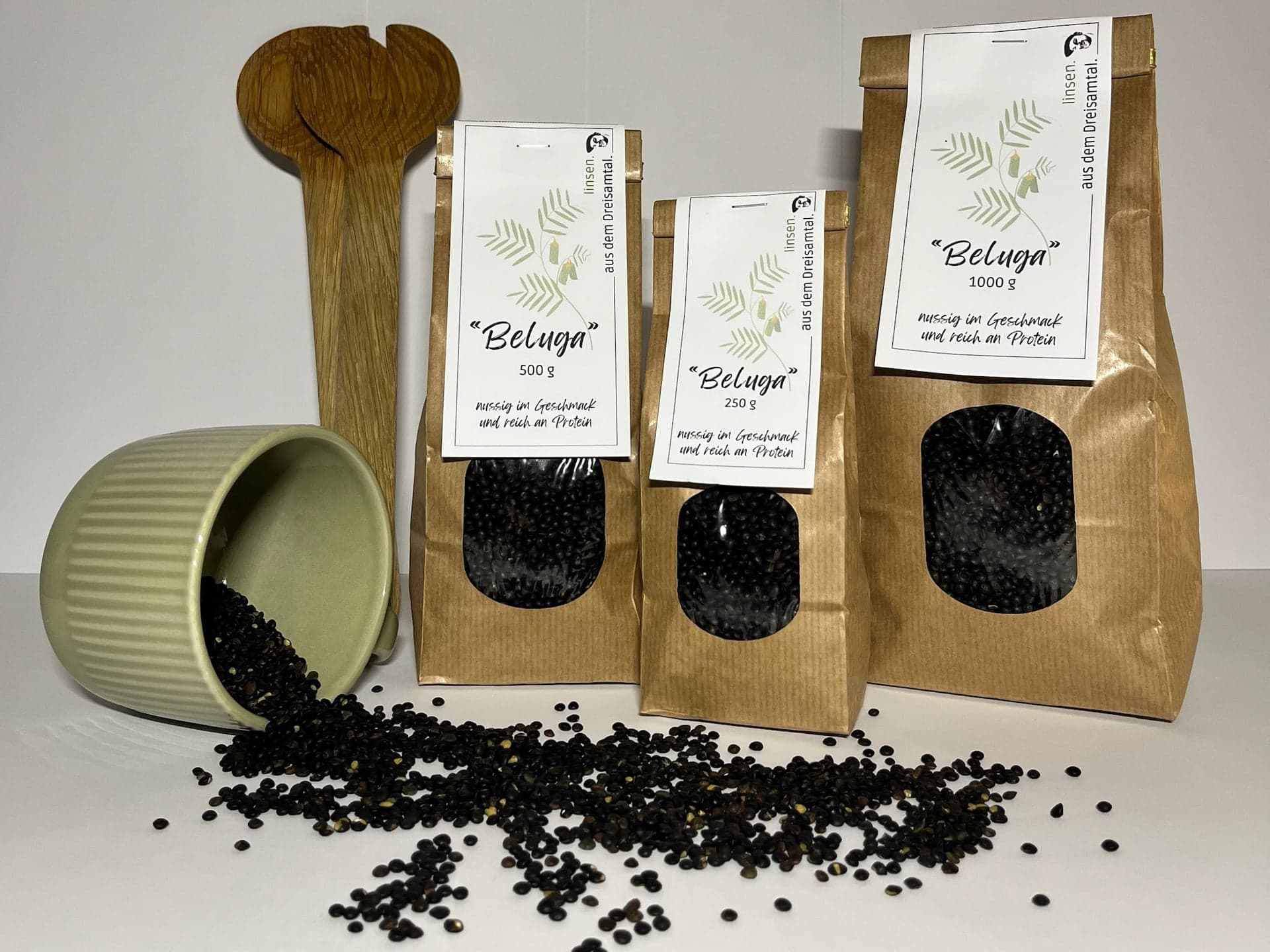 Drei braune Tüten Beluga-Linsen, 250 g, 500 g und 1 kg, daneben eine Schale schwarzer Linsen
